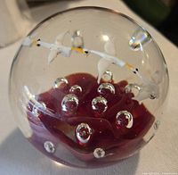 Full view of spherical glass paperweight showing red floral core, white bird figures, yellow branch, and air bubbles