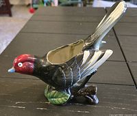 Left side view of ceramic bird planter showing red head, black body, opening on back, branch and green leaf base