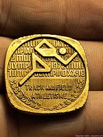 Front of square gold-tone medallion showing stylized runner, Olympiad XX 1972 text and Track and Field wording