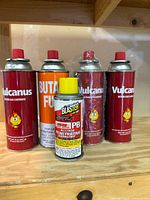 Group of four red Vulcanus butane cartridges, one orange butane cartridge, and one yellow-capped Blaster PB can on wooden shelf