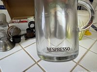 Nespresso glass mug, stainless steel funnels, black plastic adapter, box with coffee filters and accessories on tile countertop