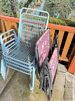 Stack of light blue metal wire patio chairs and two folded black frame floral sling chairs on patio