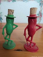 Front view of both red and green corn man bottle stoppers showing cork hats and canes