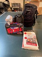 Complete kit laid out: boxed camera, lens wrapped in plastic, Canon backpack, SanDisk card pack in foreground