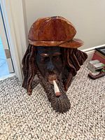 Front view of carved wooden bust showing cap, dreadlocks, cigarette