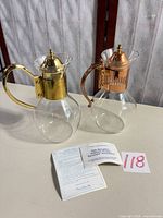 Front view of both glass carafes showing brass-tone and copper-tone tops, with paperwork