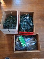 Three cardboard boxes filled with tangled green-wire Christmas string lights; front box also holds a retail carton and plastic bagged strands