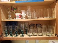 Cabinet shelves holding assorted glass tumblers, goblets, lowball glasses, silver-rim glasses, milk glass mugs, Tabasco mug