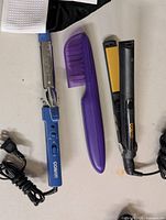 Flat lay of three hair styling items: Conair curling iron, purple comb guard, Conair straightener