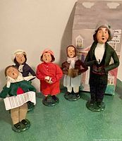 Five caroler figurines on green surface in front of house backdrop