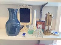 Shelf view showing blue ceramic pitcher, ornate frame, milk glass vase, glass candle holders, brass miner's lantern, small dish