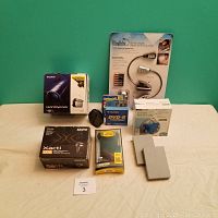 Overview of all lot items, showing boxed Sony Handycam, Sanyo Xacti CG9 camera box, UltraBrite LED booklight, Maxell batteries, Fujifilm DVD-R discs, OtterBox Defender phone case, bathtub radio, and two gray plastic items.