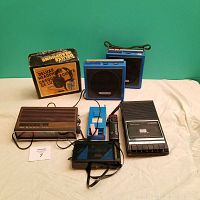 Lot arranged on a white sheet with green background, showing all seven electronic items including headphones in box, two blue speakers, cassette player, radio, handheld game, and remote control.