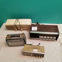 Four non-working vintage radios shown from front with all control knobs and dials visible, placed on beige cloth background, against green wall.