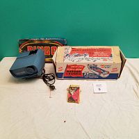 Photo showing blue ViewMaster projector with power cord, original Ideal Boat toy box, bingo game box, and small pack of cards on beige surface against green background.