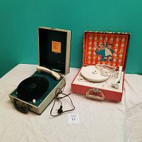 Pair of vintage portable record players side by side on fabric surface against green wall