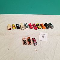 Wide shot showing 14 toy cars lined up on a white fabric background. Cars vary in size, model and color.