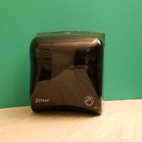 Front view of the glossy black Titan paper towel dispenser against a green background, showing the brand name and hands-free operation icon on the front panel.