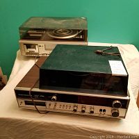 Electrohome stereo unit in front with clear dust cover on turntable, cassette deck, and 8-track player visible with control knobs on front panel; Zenith stereo unit behind it with cord on top.