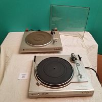 Pair of Dual turntables displayed on a white surface against a teal background. Both turntables are visible with the CS607 in front and the CS514 behind, with the dust cover set behind the CS607.