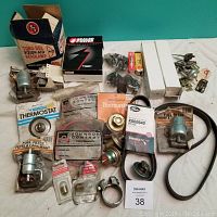 Lot of assorted vintage new auto parts arranged on a white cloth. Includes thermostats, fuel filters in blister packs, main bearing set in sealed plastic bag, bulbs in small clear packages, a black V-belt, and a Tung-Sol Vision-Aid headlamp box.