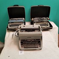 Three typewriters on a cloth-covered surface against turquoise wall. Two Smith-Corona Electra in cases on top row behind one Underwood manual typewriter in front.