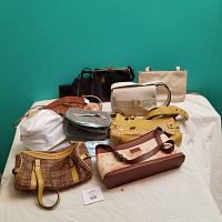 Full view of the lot showing nine purses and handbags in a variety of colors and styles arranged on white cloth surface against teal wall.