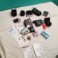 Photo showing assorted camera flashes, lenses, vintage film camera, various camera accessories, manuals, and paperwork laid out on a white cloth.