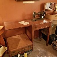 Seamstress Reversew sewing machine mounted in a wooden table with drawer and extendable side panel. Vintage brown leather carrying case underneath the table. Instruction manual on tabletop. Missing plate on machine.