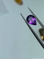 Pear-cut purple amethyst held by tweezers, showing front facet pattern