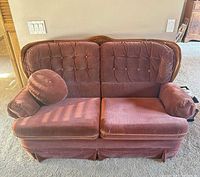 Front view of burgundy tufted loveseat showing wood trim, rolled arms, skirted base, and accent pillow