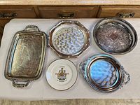 Group shot of five items: four silver-plated trays and one Centennial plate