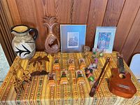 Full lot on table showing pitcher, pineapple carving, prints, figures, glasses, ukulele, flute, wood decor