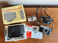 Group shot of all three primary items: Konica camera with case, Kodak Instamatic 104, Safe House alarm with box, manual and hardware
