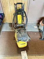 Full view of Kärcher yellow pressure washer upright with hose on handle and manual on floor