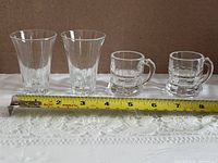Two flared faceted shot glasses and two handled mini tankard shot glasses beside measuring tape for scale
