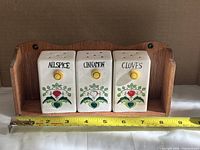 Front view of three labeled ceramic shakers seated in wood rack with measuring tape for scale