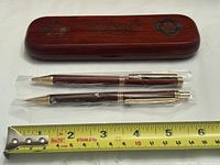 Pen and pencil in plastic sleeves beside engraved wooden case with measuring tape for scale