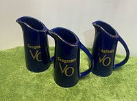 Front view of three identical blue Seagram's VO ceramic pitchers on green cloth