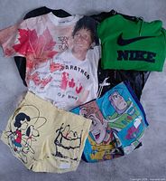Group of highlighted items: AOP Terry Fox T-shirt, green Nike hoodie, Peanuts shorts, Toy Story shorts with black bag of additional clothes