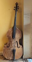 Full view of wooden double bass leaning against wall; illustrates missing bridge, fingerboard, tailpiece, body wear