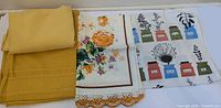 Folded mustard tablecloth, floral lace tea towel, herb print tea towel side by side