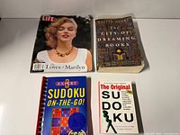 Four printed items laid out: Life Marilyn issue, City of Dreaming Books novel, Expert Sudoku spiral book, Original Sudoku paperback