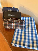 Three folded roll-up picnic blankets stacked beside folded blue gingham tablecloth
