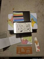 Open metal tin showing sticky tabs and colouring card; surrounding items laid out including pencil pack, ruler-stencil, stickers