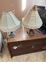 Pair of metal-base table lamps with cream fabric fringe shades on wooden surface