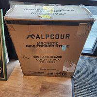 Sealed Alpcour Magnetic Bike Trainer Stand shipping carton showing branding, SKU APC-BTSMBK and color black