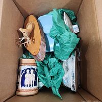 Box showing brass shell, beer stein, blue dish, deer plaque, white plaque