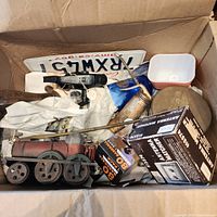 Mixed items in box including toy train car, license plate, brass sprayer, gauges, pump handle visible