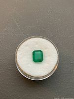 Loose rectangular emerald-cut emerald in clear round case on white cushion